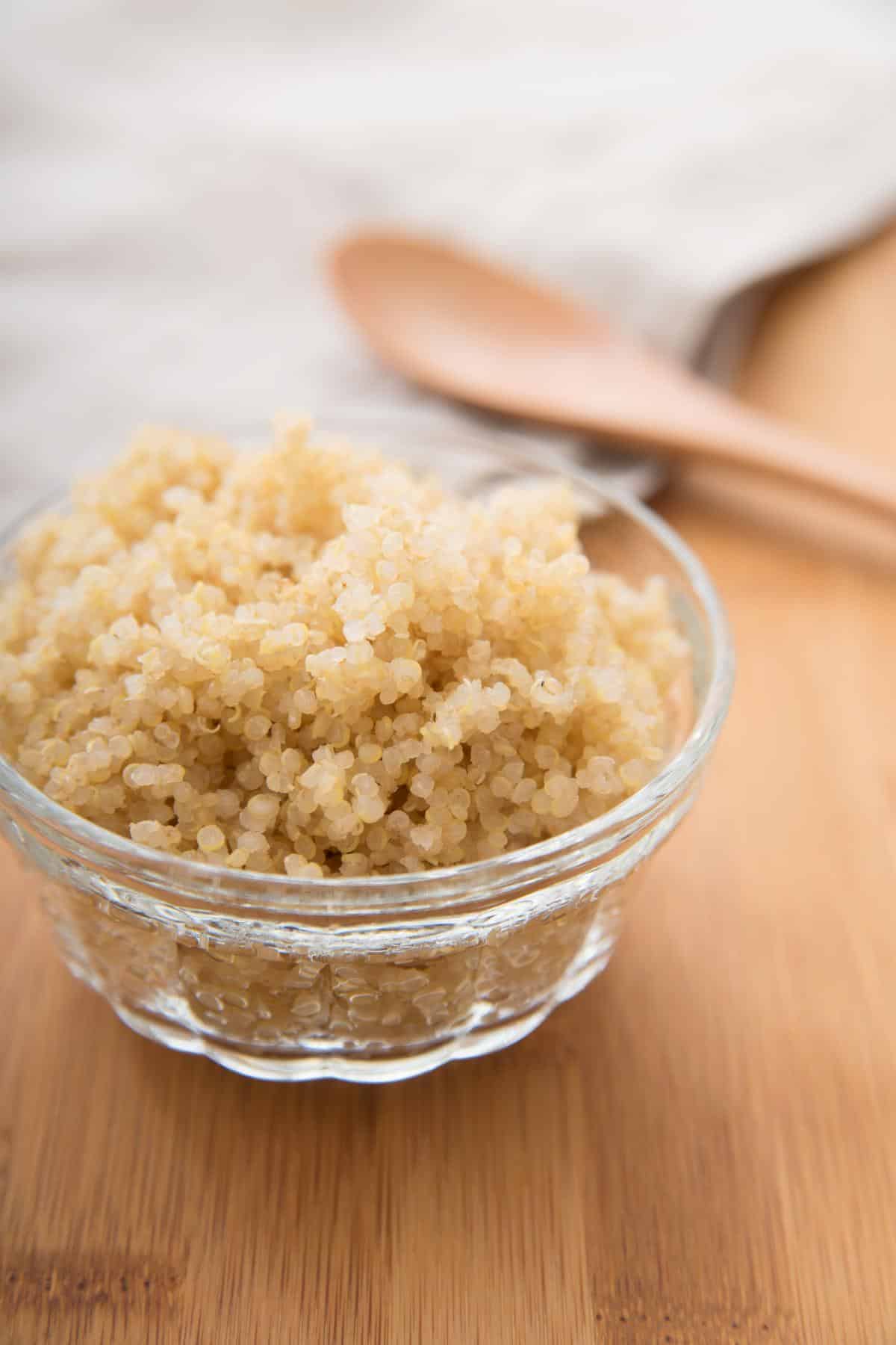 Small glass bowl with cooked white quinoa and wooden spoon in background.