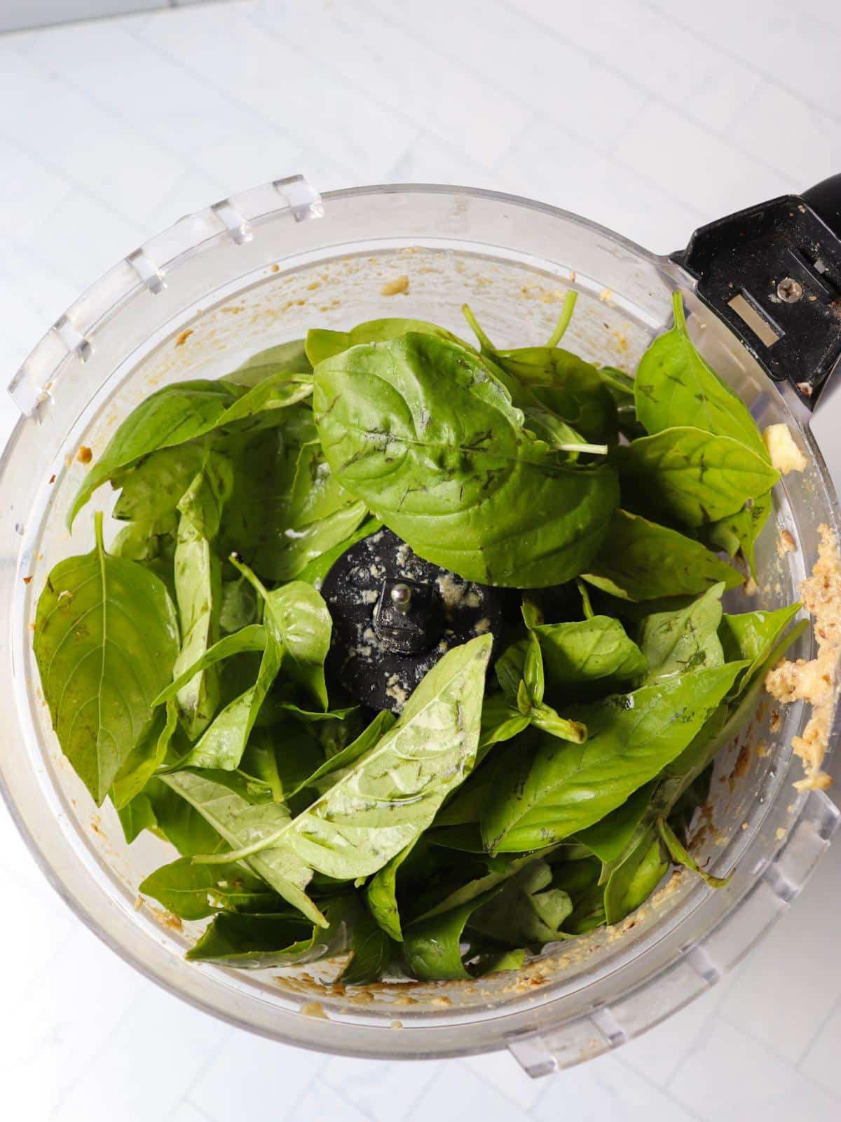 Fresh basil leaves added to a food processor with blended nuts and seasonings for vegan pesto.