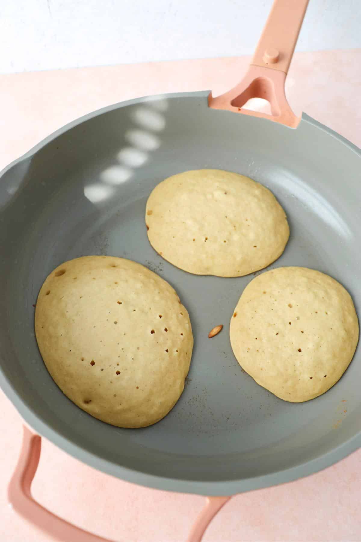 Vegan protein pancakes cooking in a skillet with bubbles on top of the pancakes.