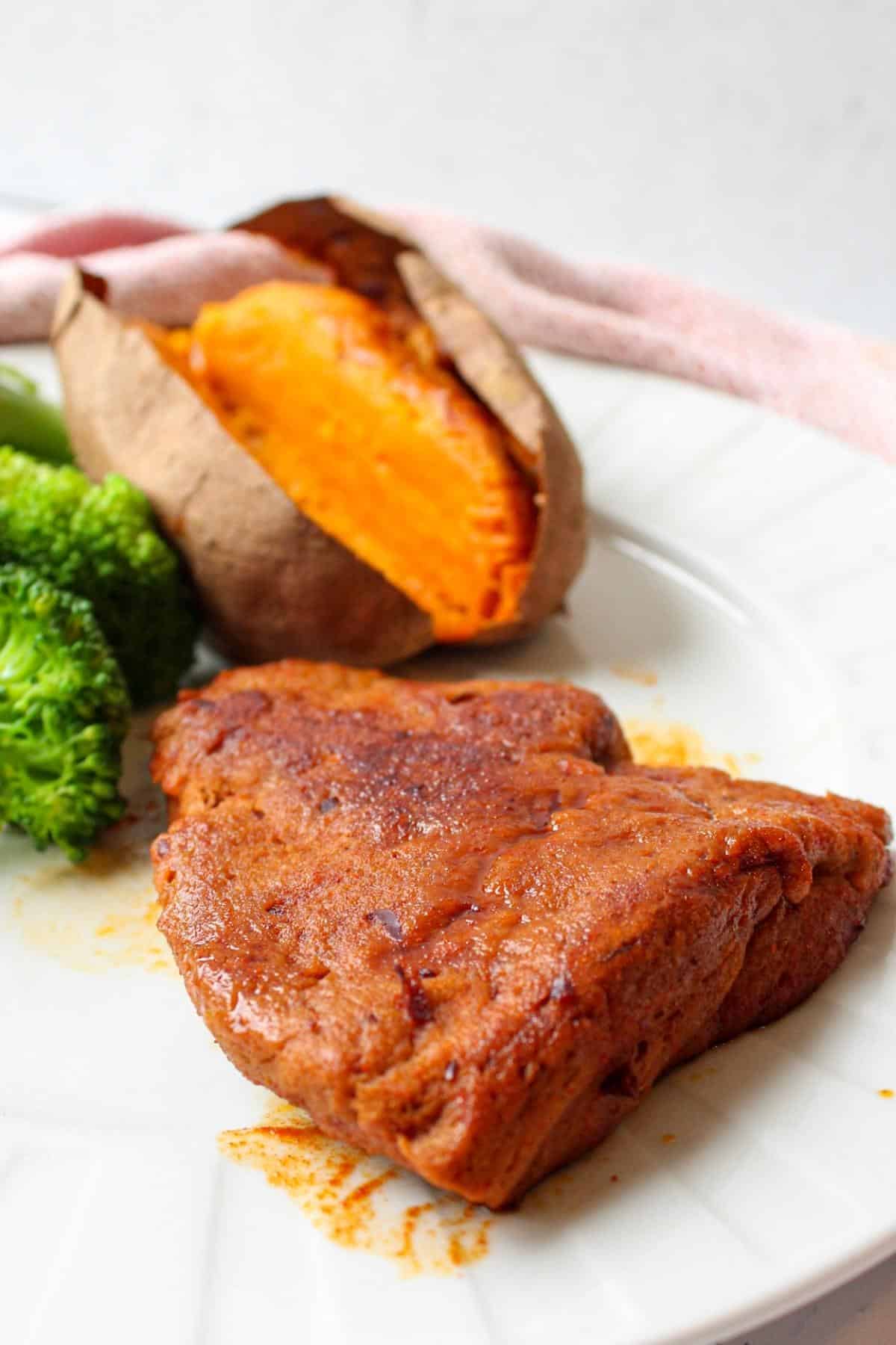 Piece of vegan steak on a plate with broccoli and a baked sweet potato.