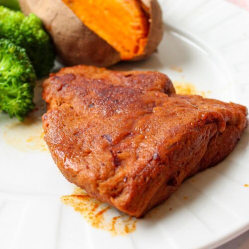 Piece of vegan steak on a plate with broccoli and a baked sweet potato.