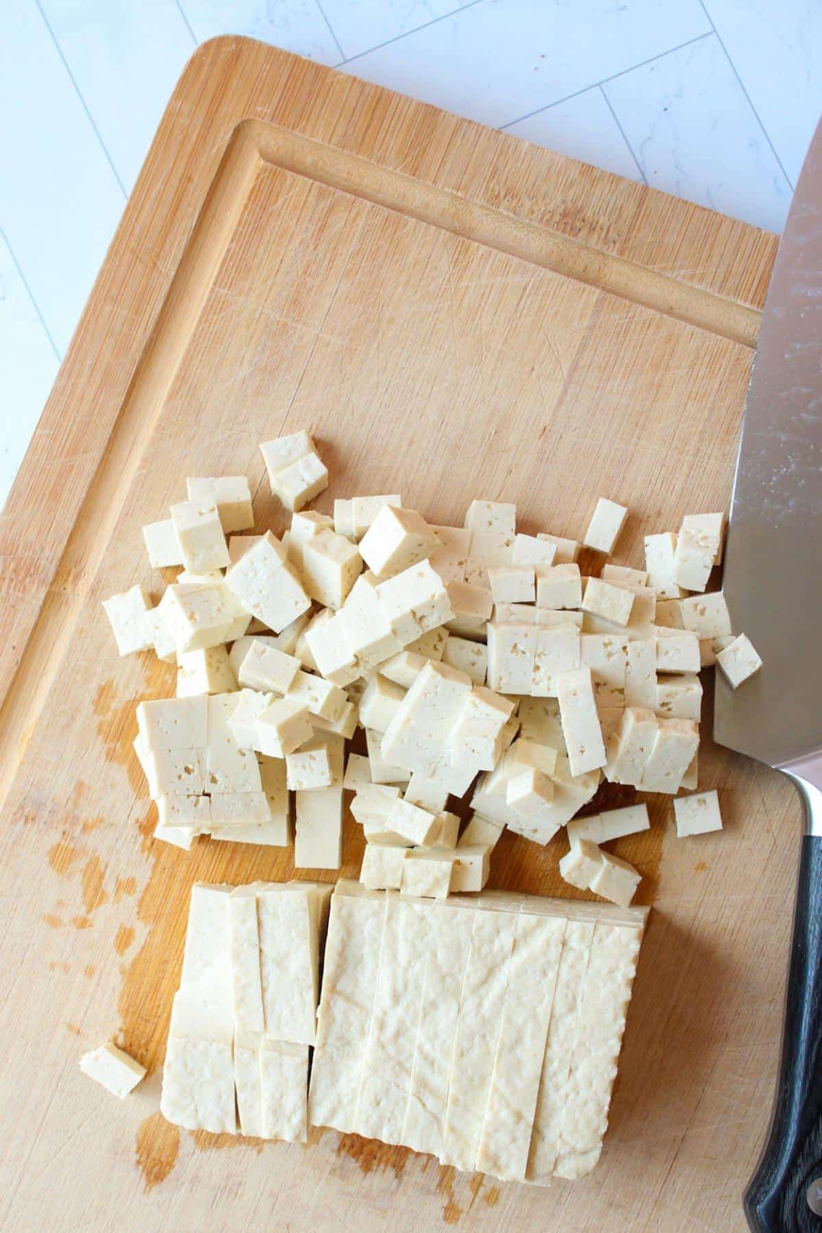 Block of tofu on a cutting board getting cut up into little cubes.