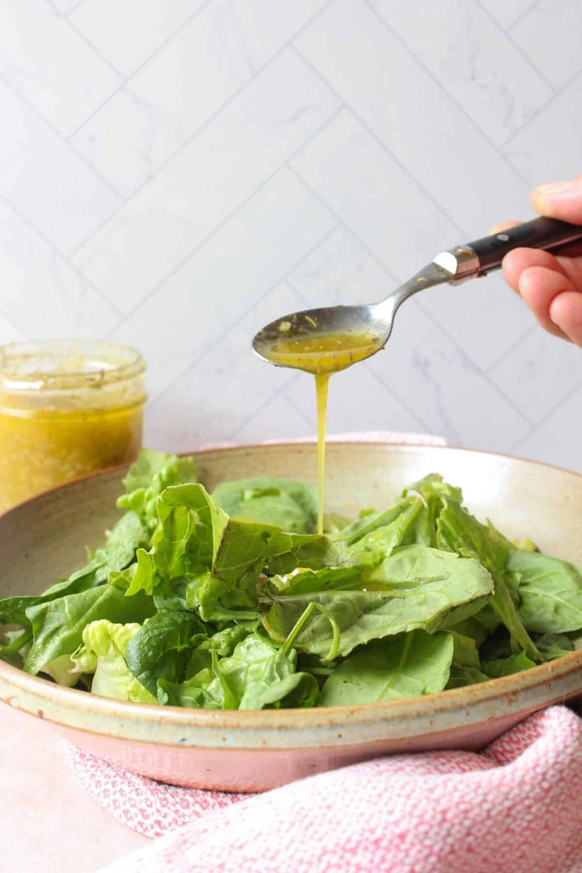 A spoon drizzling vegan Italian dressing over a bowl of lettuce leaves.