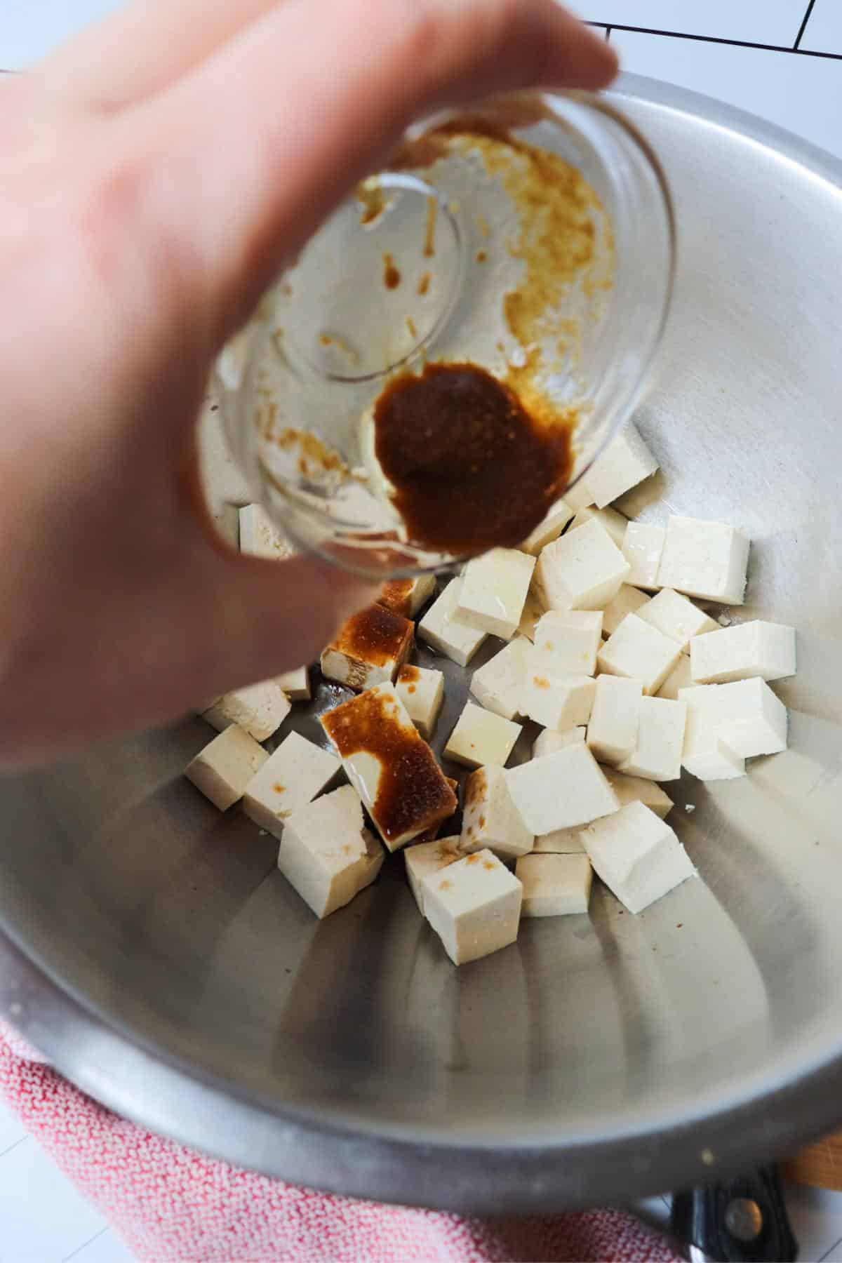 Tofu cubes in a mixing bowl with soy sauce getting drizzled on them.