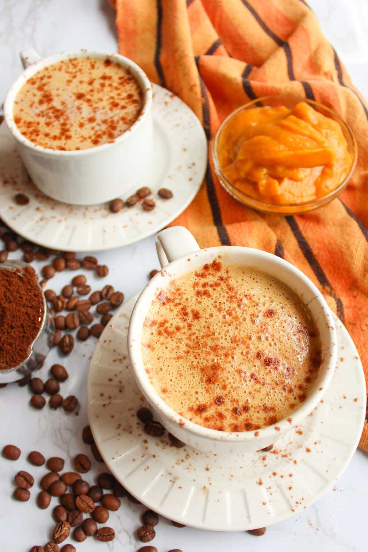 Two mugs of vegan pumpkin spice latte on a table with coffee beans and pumpkin puree.