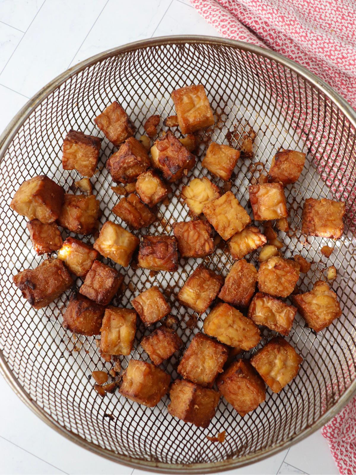 Marinated and air fried tempeh cubes in an air fryer basket on a kitchen counter next to a red kitchen towel.