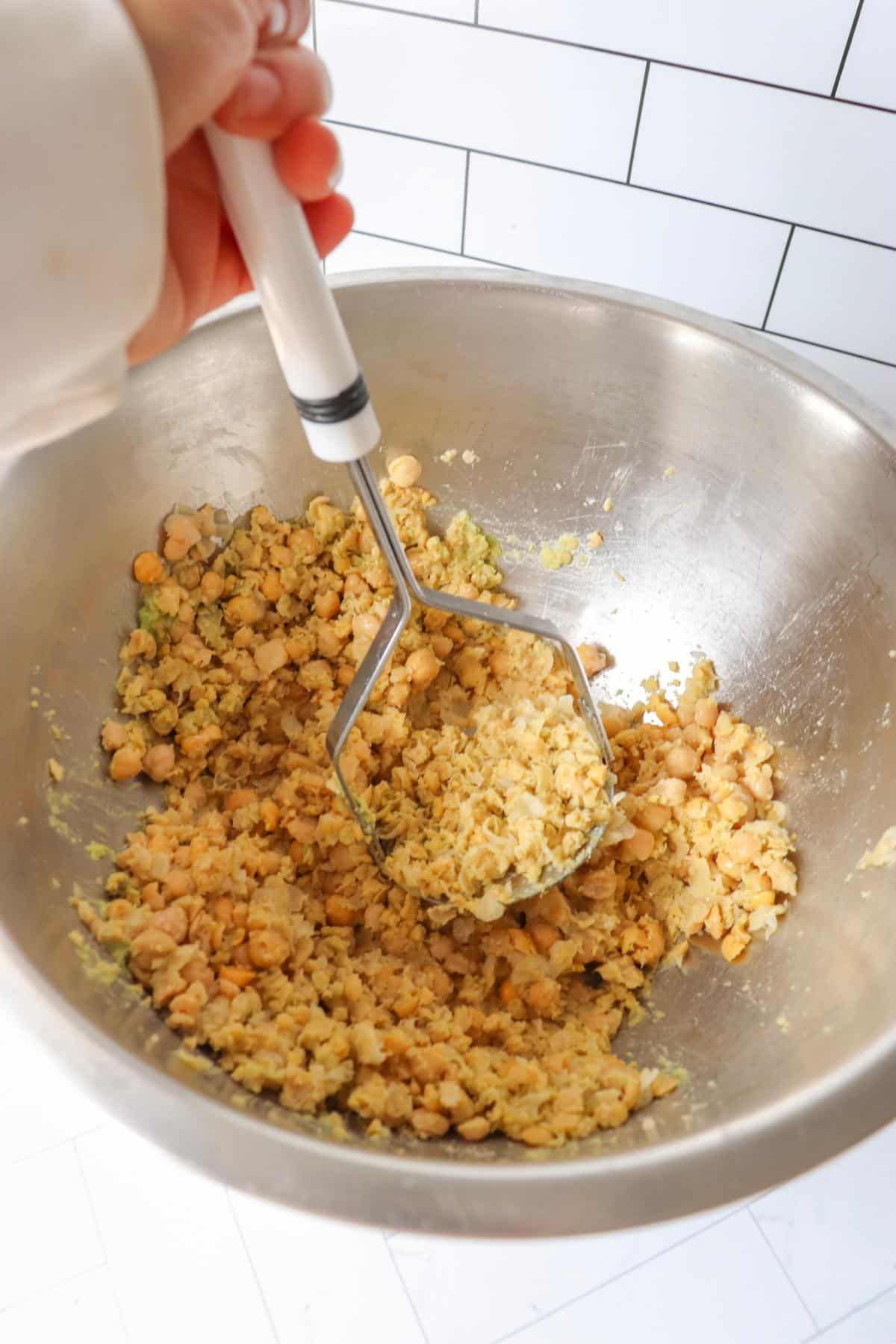 Hand using a potato masher to mash chickpeas in a large mixing bowl.