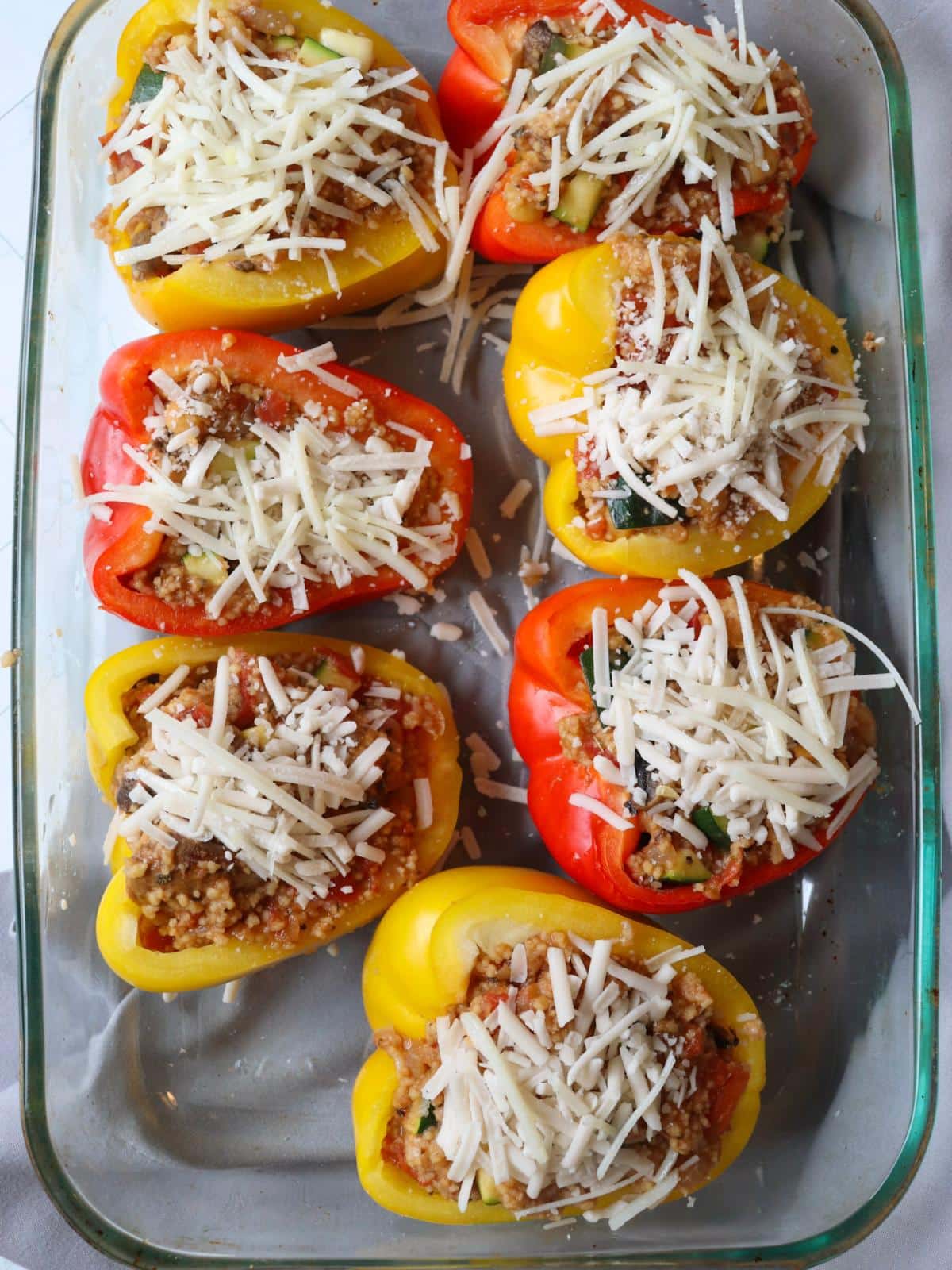 Couscous stuffed peppers in a baking dish, topped with vegan cheese, before getting roasted.