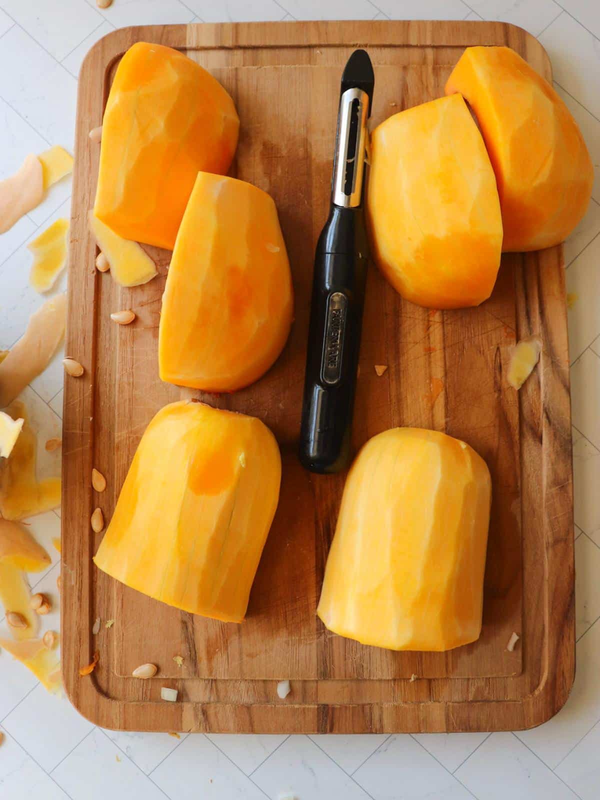 Butternut squash pieces getting peeled on a wooden cutting board.