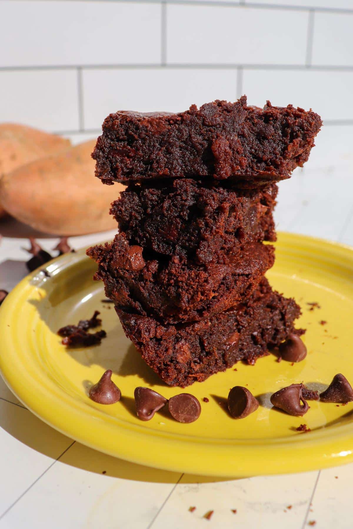 Four sweet potato brownies stacked on each other on a yellow plate with two sweet potatoes in the background.