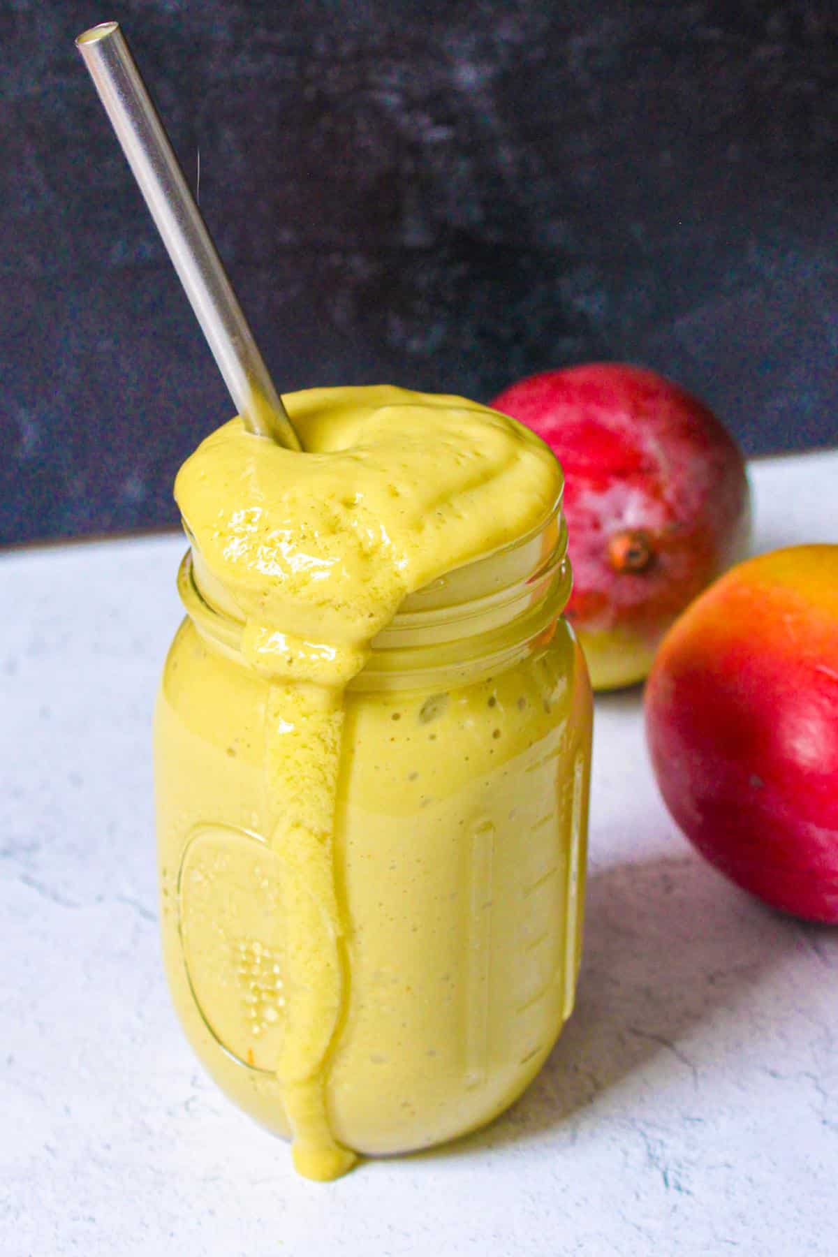 High protein mango smoothie in a glass mason jar with a metal straw and mangoes behind it.