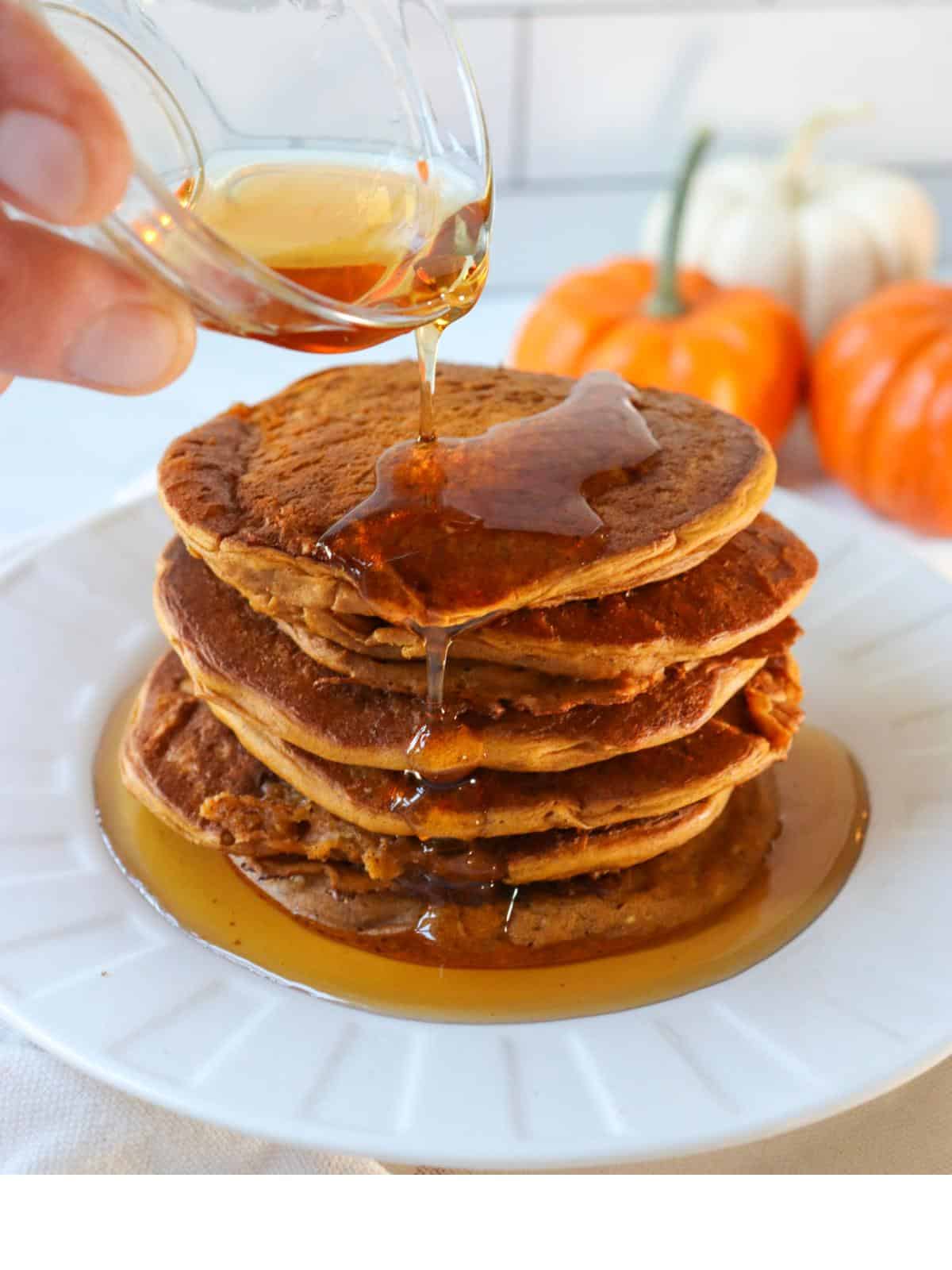 A stack of vegan pumpkin pancakes on a plate with a hand pouring maple syrup over them.