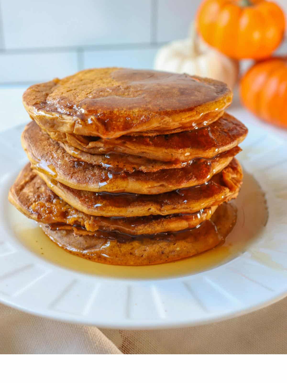 Egg free dairy free pumpkin pancakes stacked up on a white plate next to mini pumpkins.