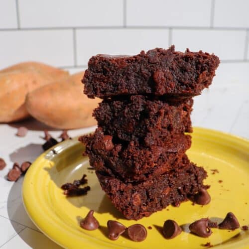 Four sweet potato brownies stacked on each other on a yellow plate with whole sweet potatoes in the background.