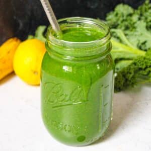 Green spirulina smoothie in a glass mason jar with a metal straw and fruit in the background.