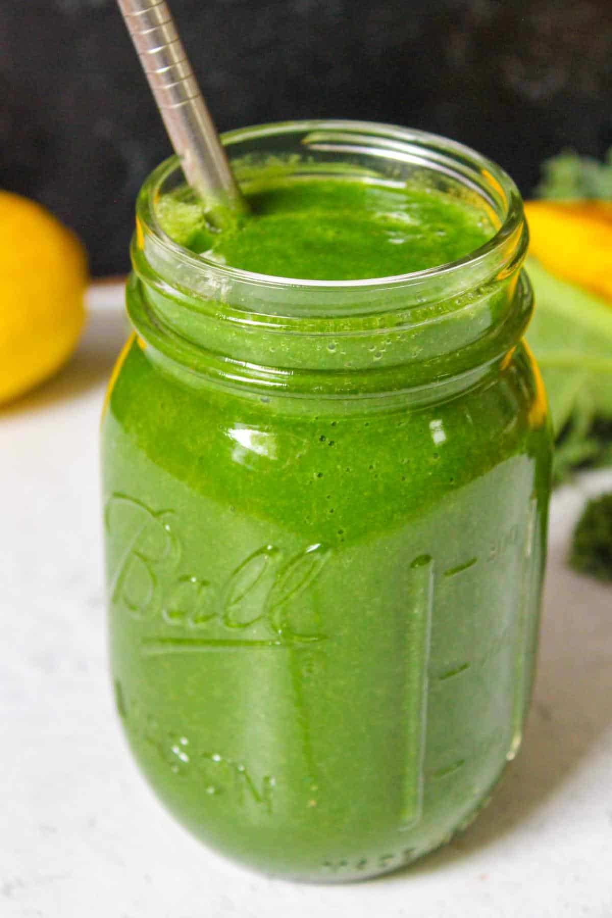 Green spirulina smoothie in a glass mason jar with a metal straw and fruit in the background.