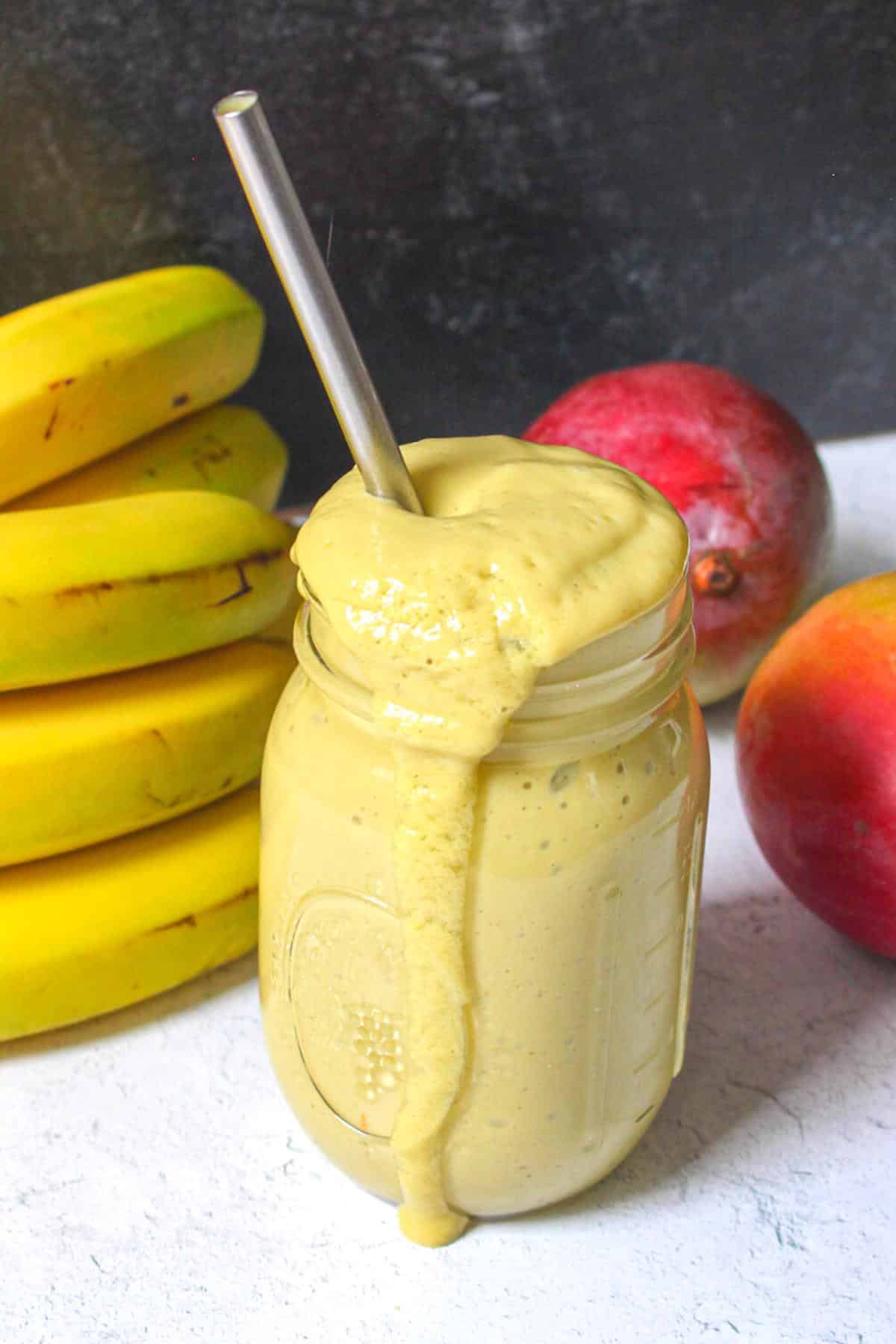 Mango protein smoothie in a glass mason jar with a metal straw with bananas and mangoes behind it.