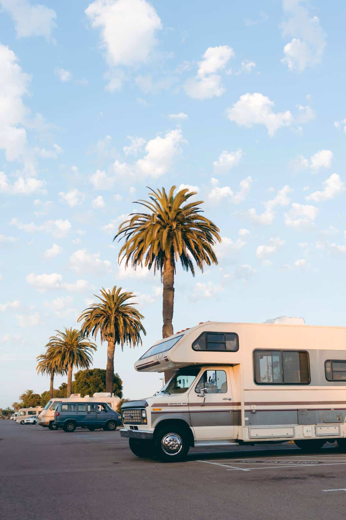 An R.V in a parking lot with a blue sky and palm trees. 