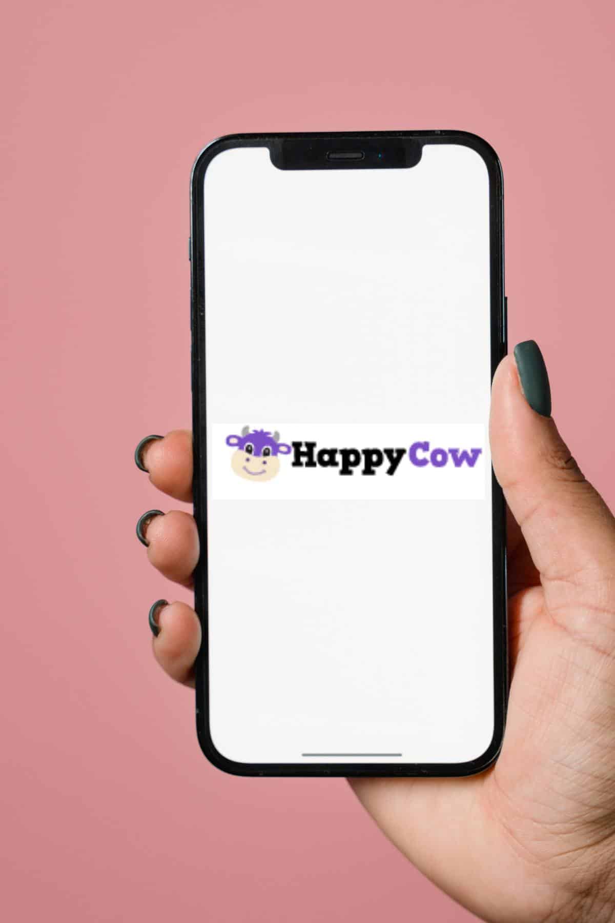 A hand holding a cellphone displaying the Happy Cow app logo. 