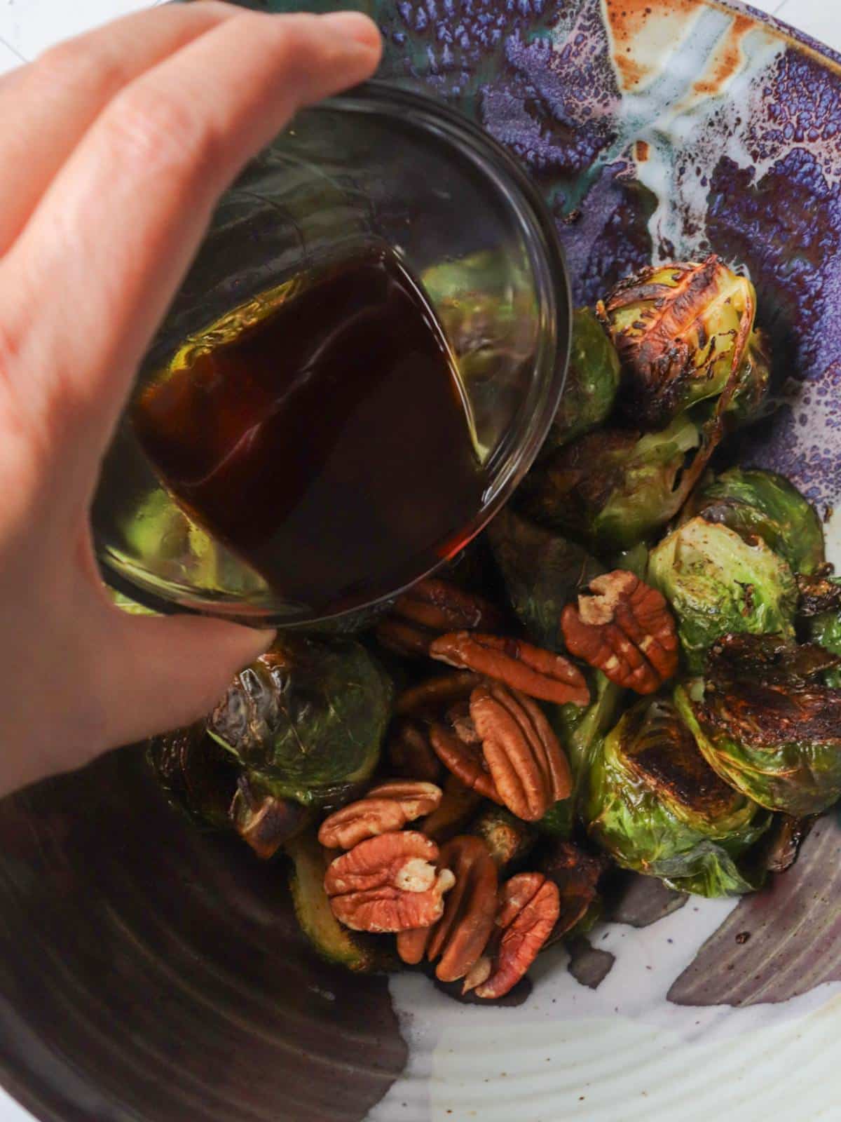 A hand pouring maple balsamic glaze over roasted brussel sprouts after being roasted.