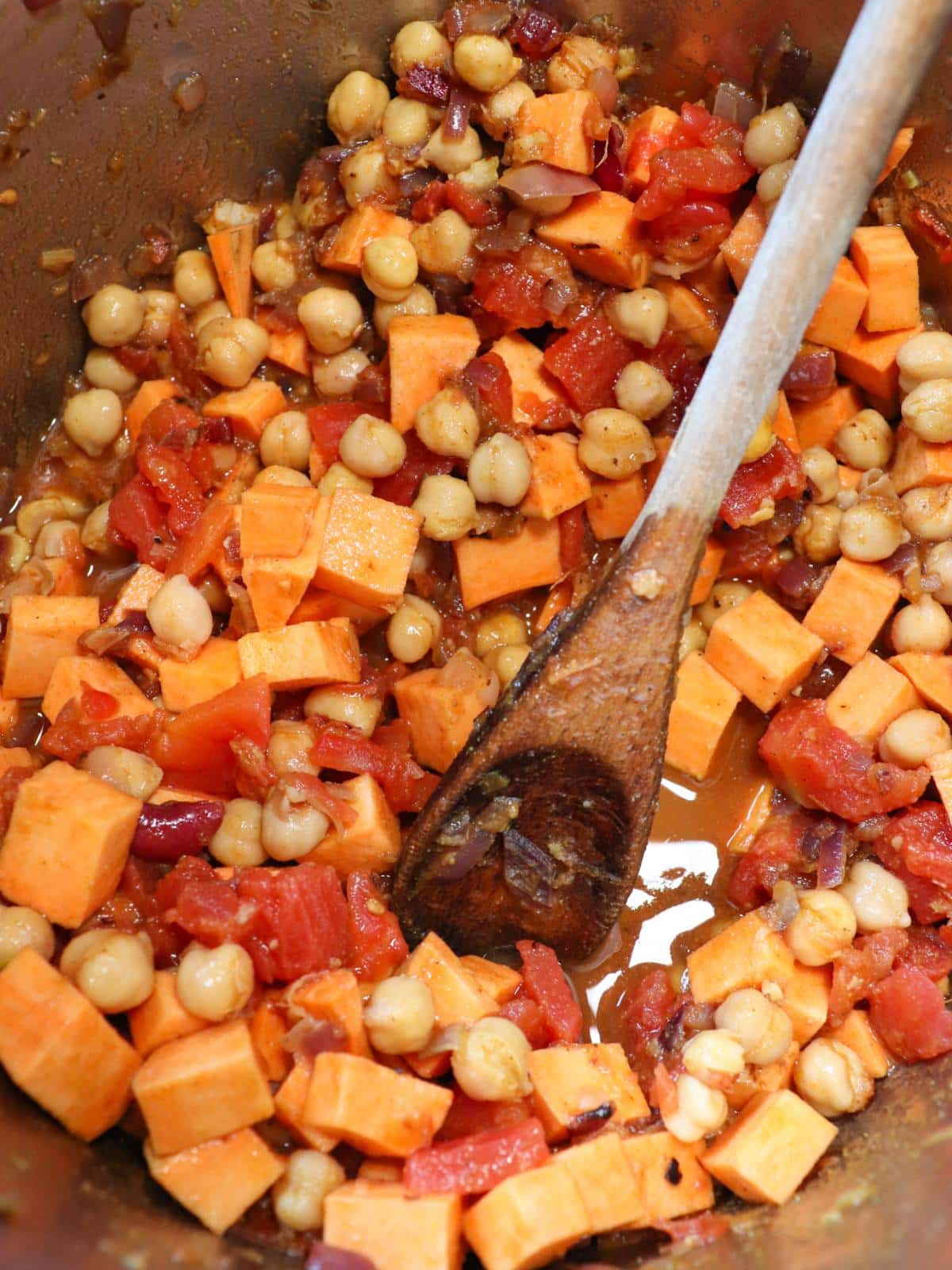 Chickpeas, sweet potatoes, tomatoes, spices, onion, garlic, ginger in an instant pot with a wooden spoon.