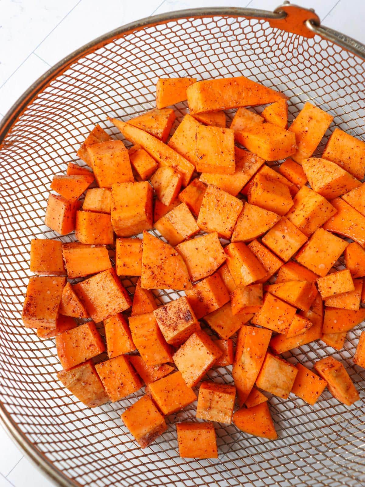 Seasoned sweet potato cubes in an air fryer basket before being air fried.