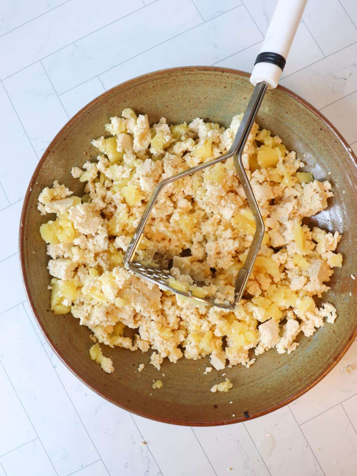 Boiled potato and crumbled tofu in a mixing bowl with a potato masher.