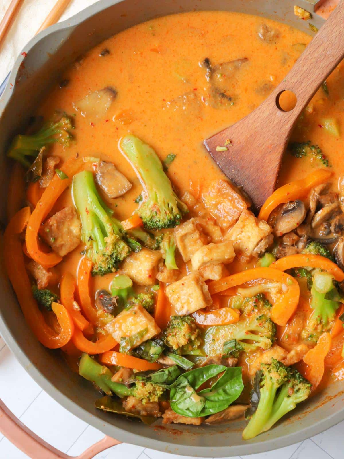 Vegan red thai curry with tofu and vegetables in a large pan with a wooden spoon.