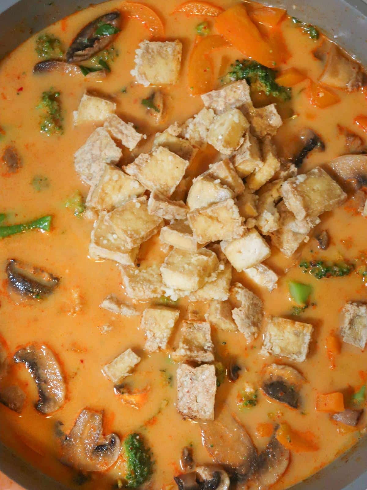 Baked tofu being added to vegan red thai curry sauce in a pan.