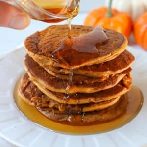 Vegan pumpkin pancakes on a white plate with maple syrup next to mini pumpkins.