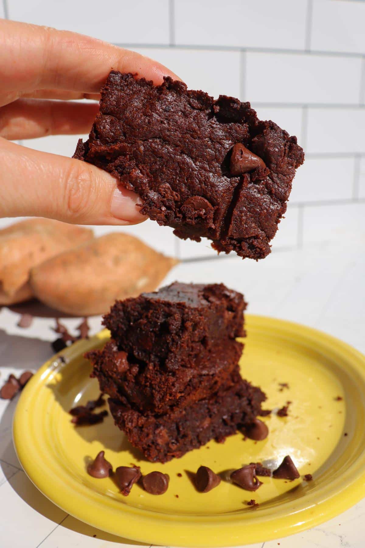 Hand holding a sweet potato brownie up in the foreground, a plate of brownies in the background.