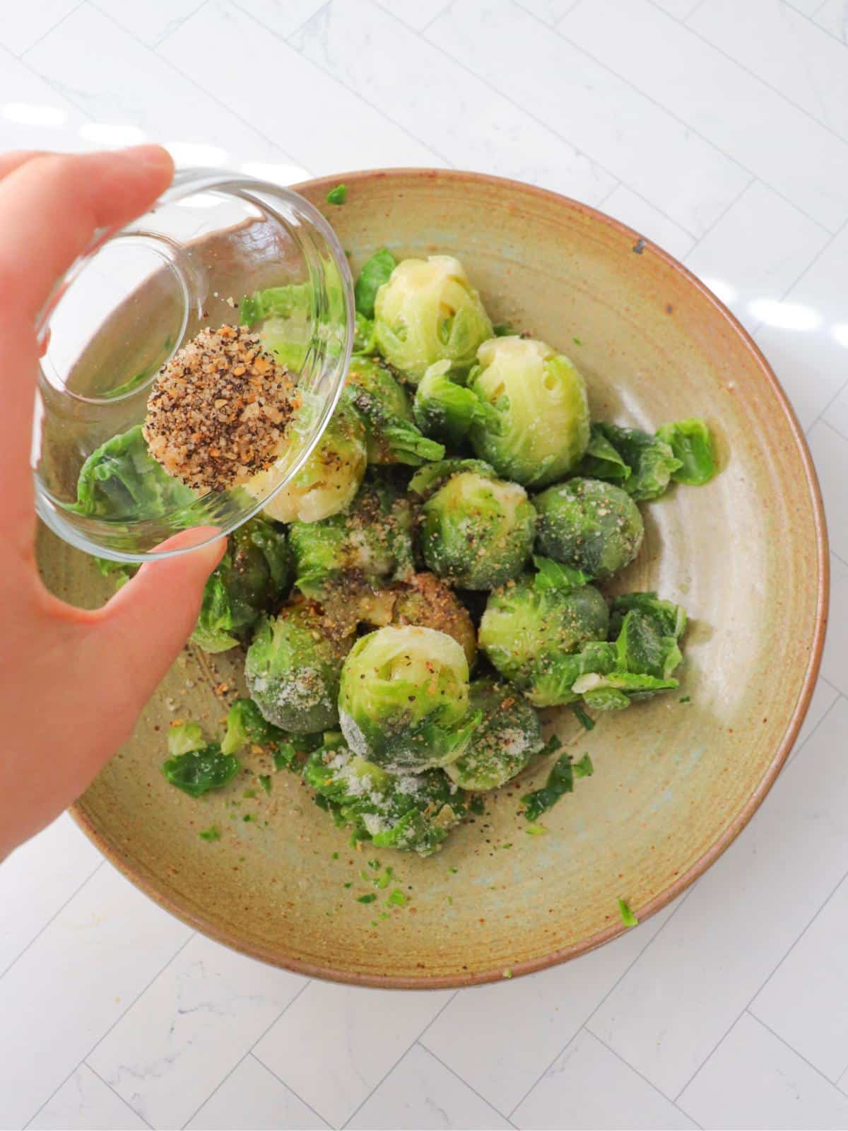 A hand sprinkling seasonings onto frozen brussels sprouts in a brown ceramic bowl.